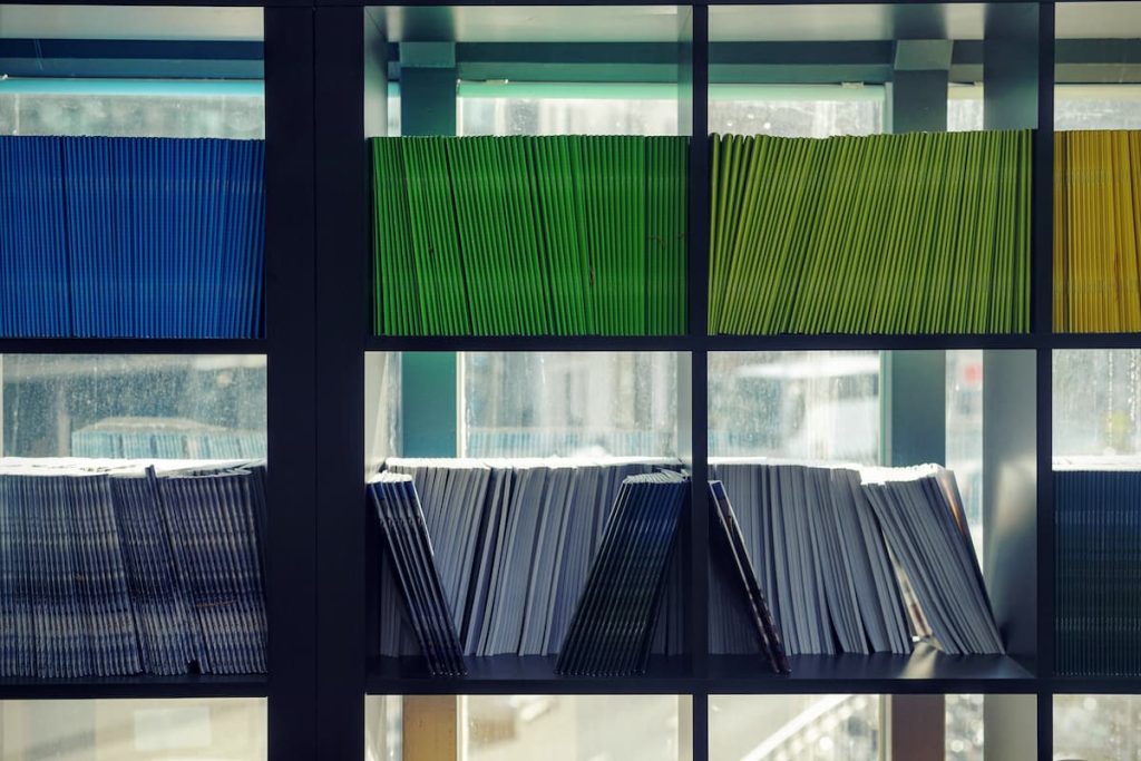 Des étagères remplies de dossiers colorés en bleu, vert, jaune et blanc, alignés et empilés, placées devant une fenêtre lumineuse.