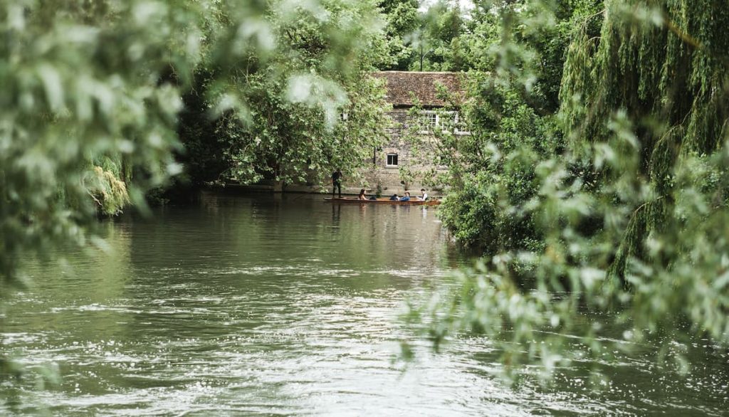 Vue d'une rivière entourée de verdure, avec des rameurs dans une barque au loin, près d'une maison en pierre partiellement cachée par les arbres.