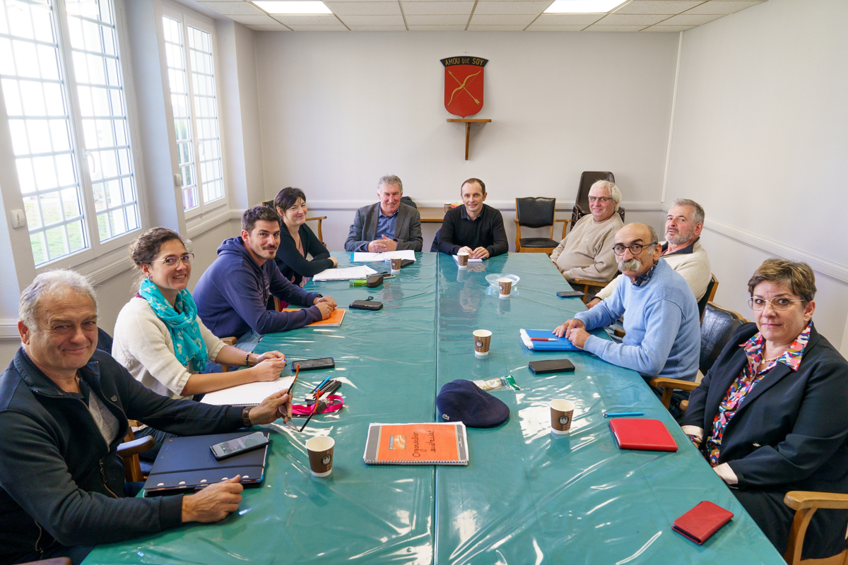 Groupe de personnes autour d'une table lors d'une réunion, dans une salle éclairée par des fenêtres, discutant et prenant des notes.
