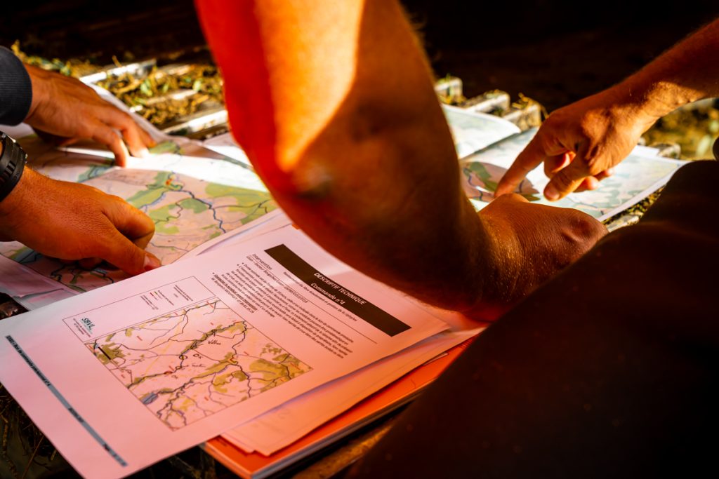 Des mains consultent des cartes topographiques et des documents imprimés, discutant d'une trajectoire ou d'un itinéraire sous une lumière chaleureuse.